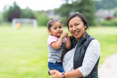 Native woman smiling and holding her toddler child with green grass out of focus in background