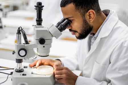 young black man in white lab coats looks through microscope in lab setting