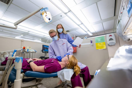 A student practices dental hygiene techniques on another student while a faculty member observes.