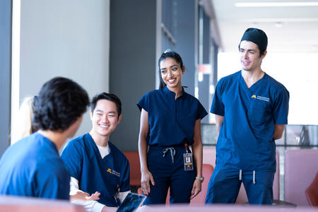 Four students in navy blue scrubs gather and talk around a lounge space in HSEC