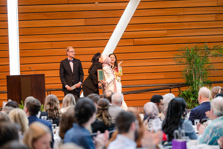 A faculty member hugs a student while two others stand on stage during an awards ceremony
