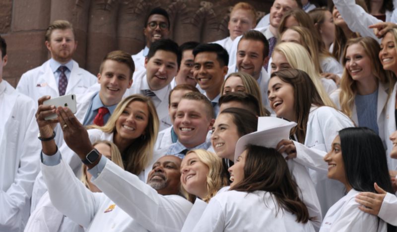 Students in white lab coats gathered on steps taking selfie