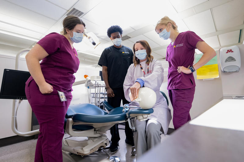 group of four dental hygiene students watch a student demonstrate on a plastic model patient