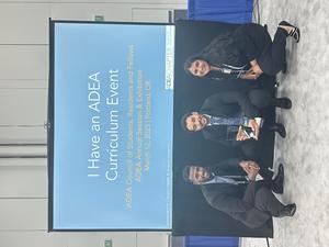 Three students pose in front of "I have an ADEA Curriculum Event" digital sign